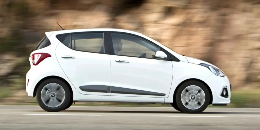 A white Hyundai i10 hatchback car driving on a winding road with a scenic landscape in the background. The car is in motion, and the background is blurred due to the speed.