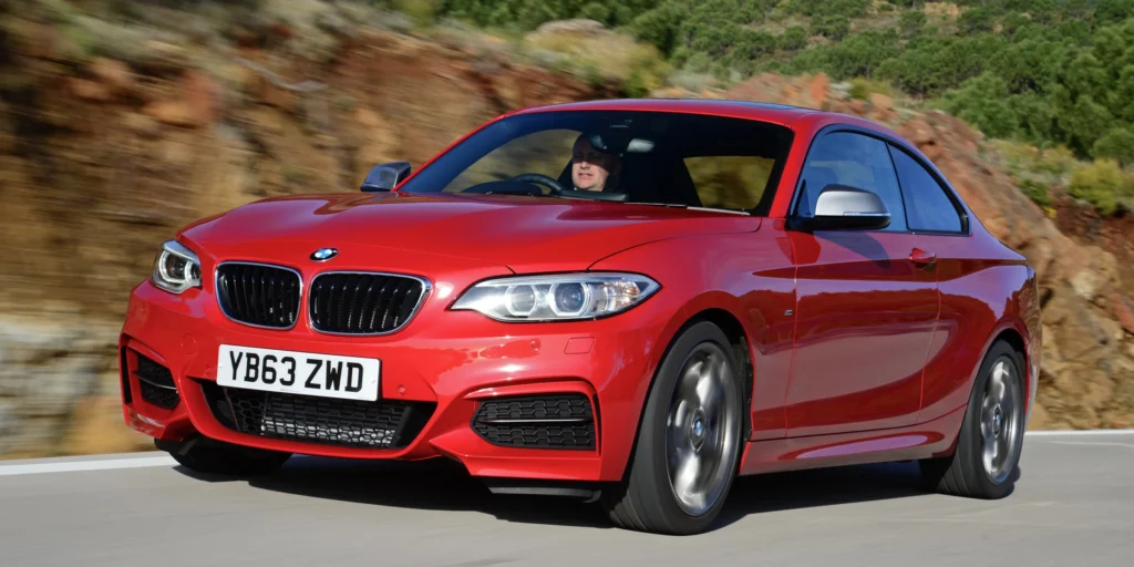 A red BMW 2 Series Coupe car driving on a winding road with a scenic landscape in the background. The car is in motion, and the background is blurred due to the speed. The license plate on the car is YB63 ZWD.