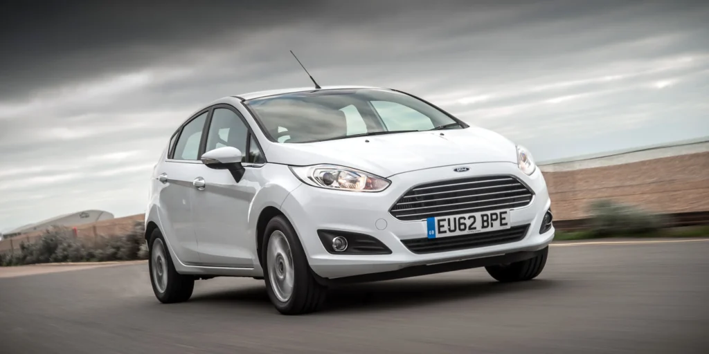 A white Ford Fiesta hatchback car driving on a road with a cloudy sky in the background. The car is in motion, and the background is blurred due to the speed. The license plate on the car is EU62 BPE.