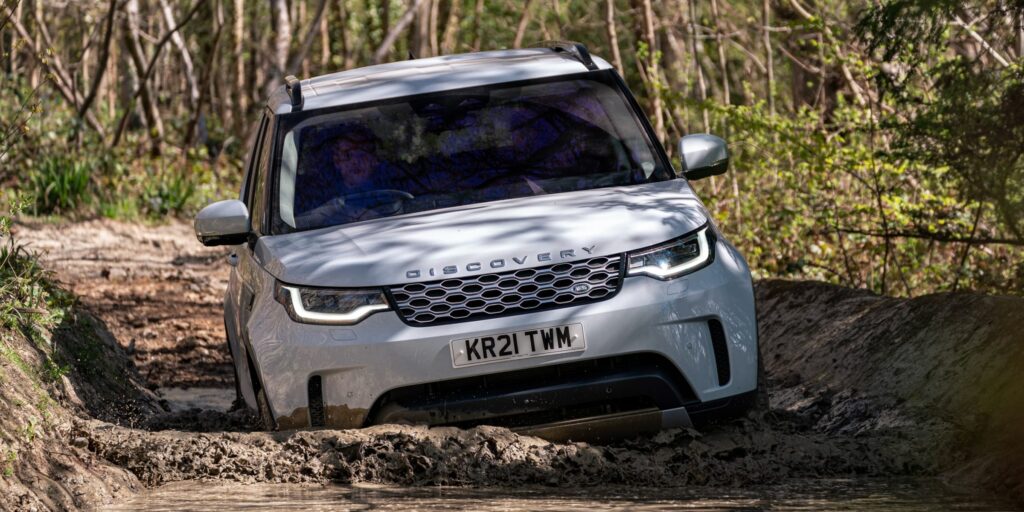 White Land Rover Discovery Sport driving through a muddy puddle on a forest trail.