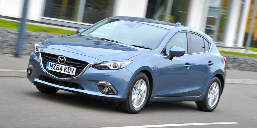 A blue 2014 Mazda3 hatchback car driving down a street. The car has alloy wheels and a modern design. The license plate on the car is MZ64 KDV.