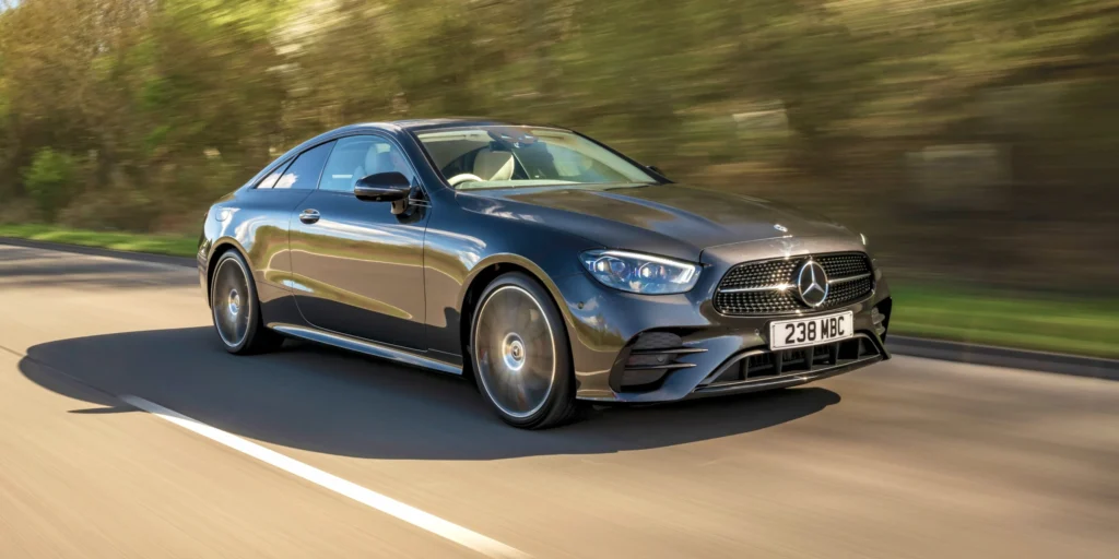 Dark gray Mercedes-Benz E-Class Coupe driving on a road with trees on either side.