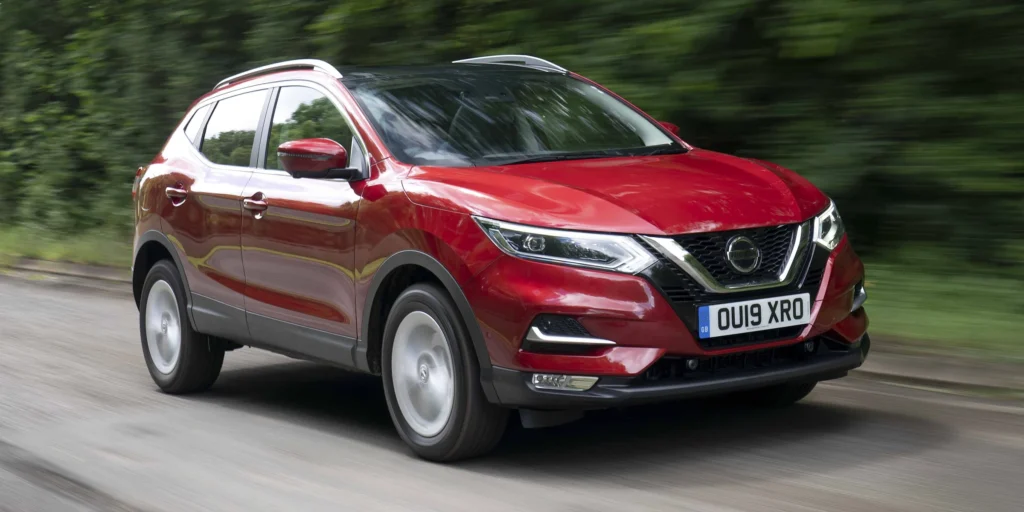 A red Nissan Qashqai SUV driving on a road with trees on both sides. The car is in motion, and the background is blurred due to the speed. The license plate on the car is OU19 XRO.