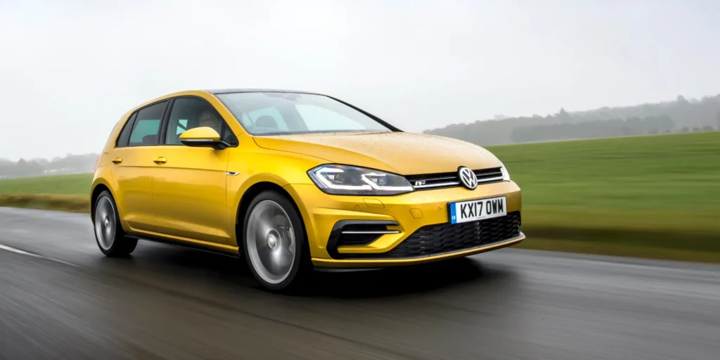 Yellow Volkswagen Golf driving on a wet road with a field in the background.