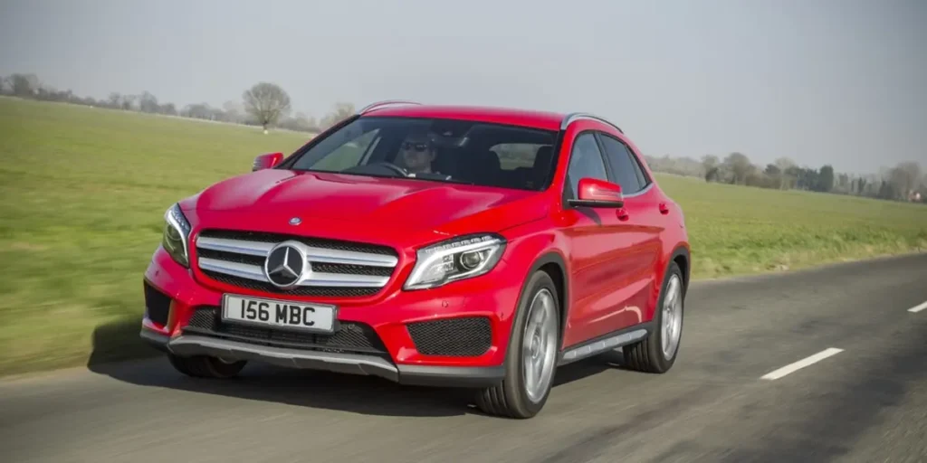 A red Mercedes-Benz GLA-Class SUV driving down a road with a scenic landscape in the background. The car has alloy wheels and a modern design. The license plate on the car is 156 MBC.