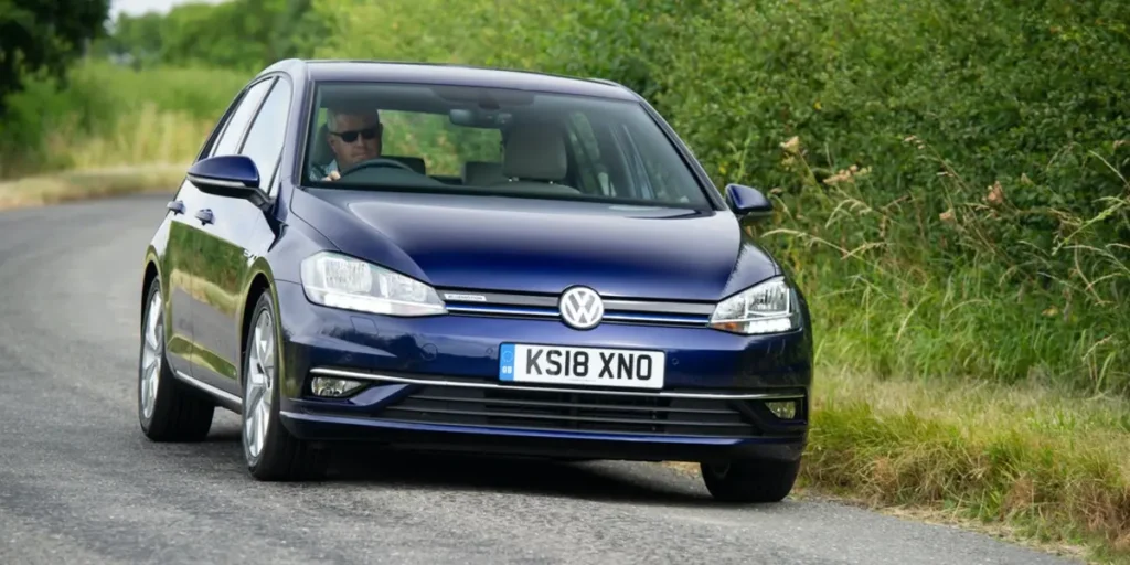 A blue Volkswagen Golf car driving on a road with a hedge on one side and a field on the other. The car is in motion, and the background is blurred due to the speed. The license plate on the car is KS18 XNO.